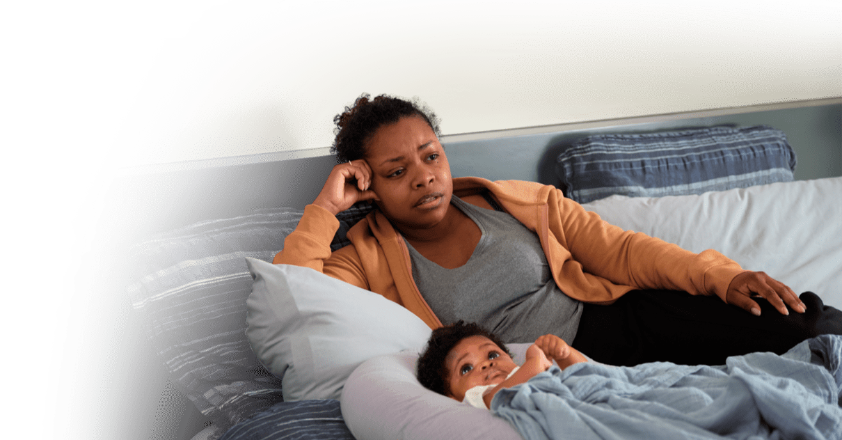 Black woman sitting on couch looking into the distance with a depressed expression and has her new baby lying next to her.